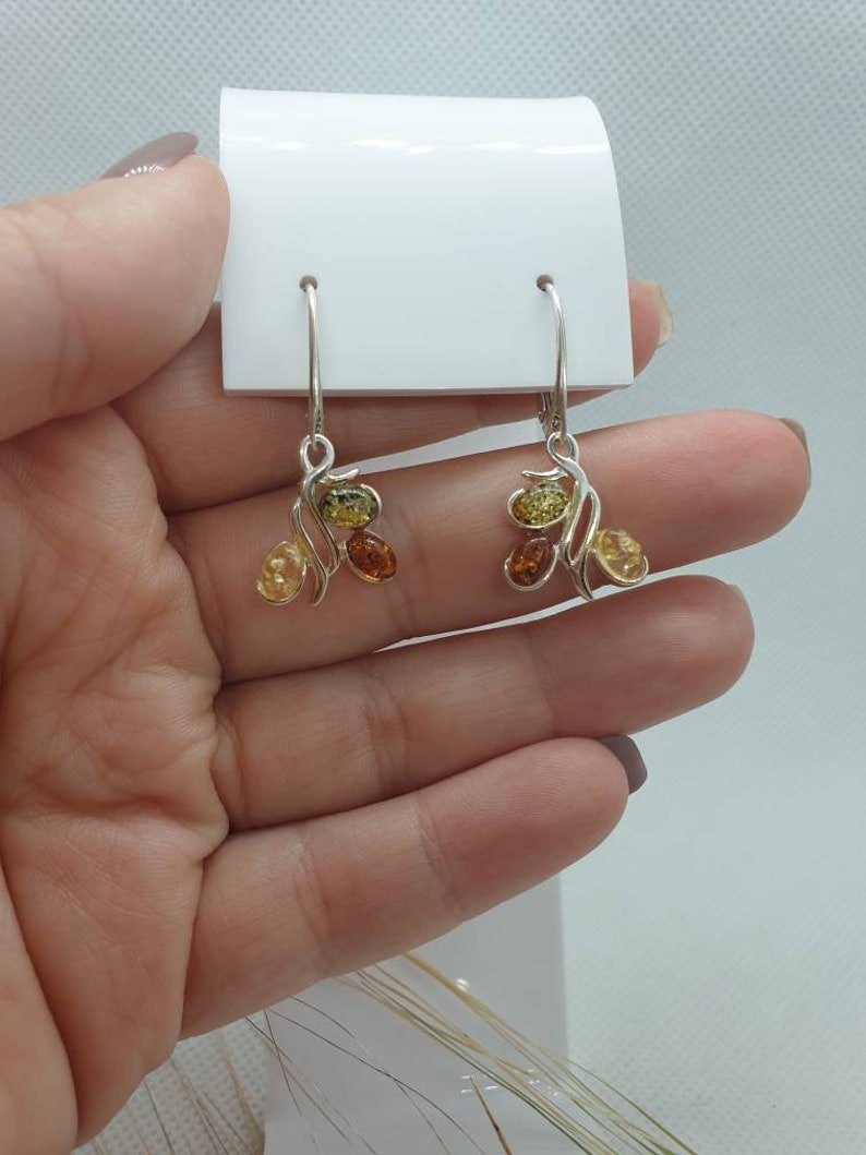 Op de afbeelding: Zilveren oorhangers met amberstenen in tinten geel, oranje en bruin. De oorhangers hebben een delicaat, gebogen ontwerp.