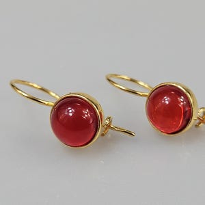 Puede incluir: Un par de pendientes dorados con una piedra preciosa roja y redonda. Los pendientes tienen un diseño sencillo con cierre de gancho. Las piedras rojas están engastadas en un bisel dorado.