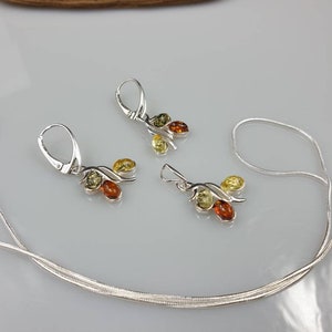Op de afbeelding: Een zilveren ketting en oorbellen set met drie amberstenen in tinten geel, groen en bruin. De ketting is een eenvoudige zilveren ketting en de oorbellen zijn van het hangende type.