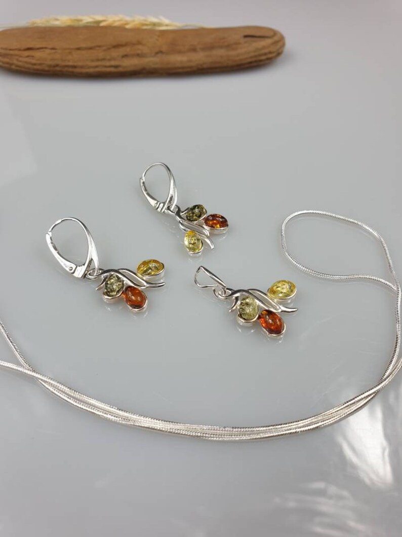 Op de afbeelding: Een zilveren ketting en oorbellen set met drie amberstenen in tinten geel, groen en bruin. De ketting is een eenvoudige zilveren ketting en de oorbellen zijn van het hangende type.