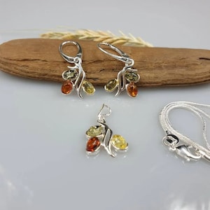 Op de afbeelding: Een zilveren ketting en oorbellen set met barnsteen stenen. De ketting heeft een hanger met drie barnsteen stenen in een bladvormig ontwerp. De oorbellen hebben een vergelijkbaar ontwerp met drie barnsteen stenen.