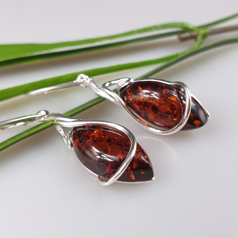 May include: A pair of silver earrings with amber teardrop-shaped stones. The stones are a deep reddish-brown color and are set in a twisted silver wire.
