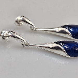 Puede incluir: Un par de pendientes colgantes plateados. Cada pendiente tiene forma de lágrima, con una piedra ovalada azul oscuro y brillante engastada en una carcasa de metal plateado. Cierre de poste y mariposa.