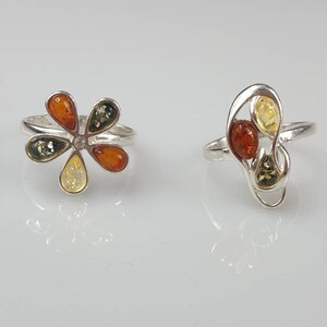 May include: Two silver rings set with amber stones. The first ring features a flower design with five amber stones in shades of green, yellow, and orange. The second ring has a more abstract design with three amber stones in shades of yellow, orange, and green.