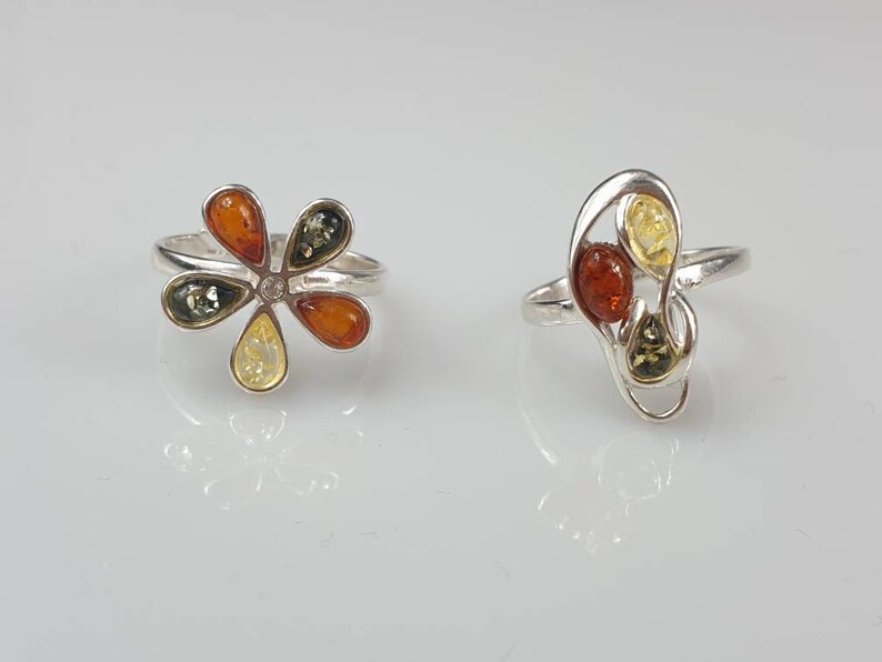 May include: Two silver rings set with amber stones. The first ring features a flower design with five amber stones in shades of green, yellow, and orange. The second ring has a more abstract design with three amber stones in shades of yellow, orange, and green.