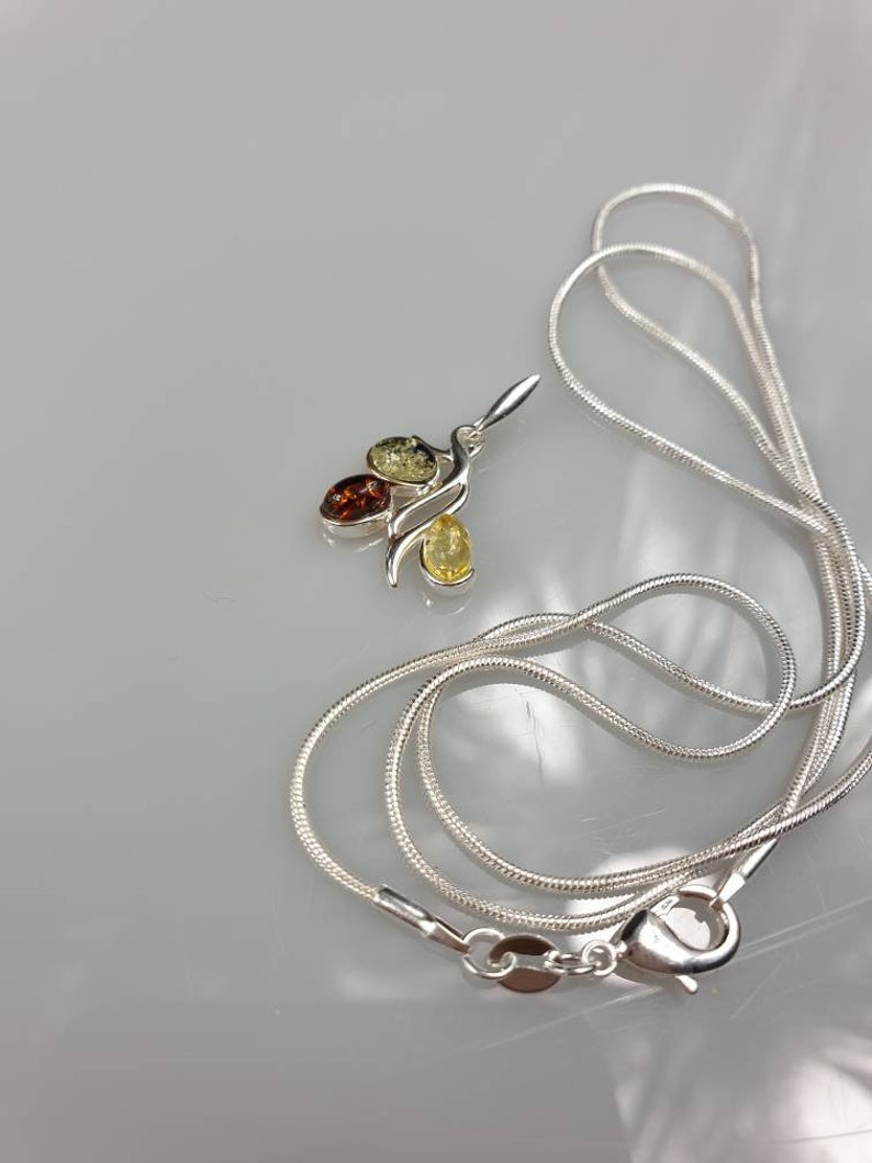 Op de afbeelding: Een zilveren ketting met een hanger met drie amberstenen in tinten geel, oranje en bruin.