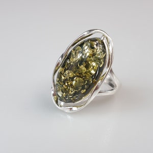 Könnte beinhalten: Silberring mit einem ovalen Bernstein-Stein in Grün- und Goldtönen. Der Bernstein ist in einen Silberrahmen mit offenem, fließendem Design gefasst. Der Ring hat ein schlichtes, poliertes Silberband. Maße: ca. 2.5 cm.