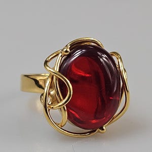Peut inclure: Une bague en or avec une grande pierre précieuse rouge de forme ovale sertie dans un délicat motif d'or en spirale.