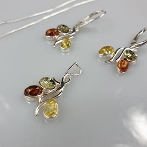 Op de afbeelding: Een zilveren ketting en oorbellen set met barnsteen stenen. De ketting en oorbellen hebben een gedraaid ontwerp met drie barnsteen stenen in elk stuk. De stenen zijn een mix van groen, geel en oranje.