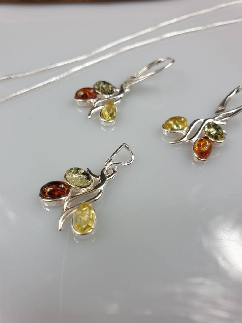 Op de afbeelding: Een zilveren ketting en oorbellen set met barnsteen stenen. De ketting en oorbellen hebben een gedraaid ontwerp met drie barnsteen stenen in elk stuk. De stenen zijn een mix van groen, geel en oranje.