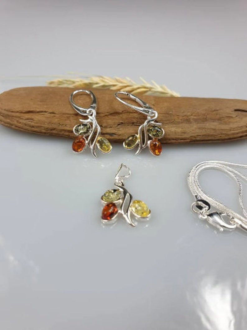 Op de afbeelding: Een zilveren ketting en oorbellen set met barnsteen stenen. De ketting heeft een hanger met drie barnsteen stenen in een bladvormig ontwerp. De oorbellen hebben een vergelijkbaar ontwerp met drie barnsteen stenen.