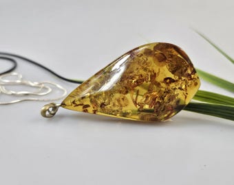 May include: A teardrop-shaped amber pendant with inclusions, suspended from a silver chain. The amber is a translucent golden-yellow color, with visible plant matter and other organic material inside. The pendant is approximately 2 inches long.