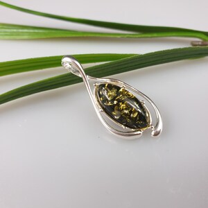May include: A silver pendant with a green amber stone set in a delicate, twisted frame. The pendant is photographed on a green leaf.