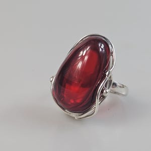 Può includere: Un anello in argento con una grande pietra rosso scuro, liscia e di forma ovale, incastonata in una montatura in metallo lucido. L'incastonatura è composta da sottili fili d'argento ricurvi che avvolgono la base della pietra.