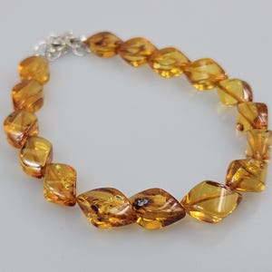 Può includere: Un braccialetto con perline di ambra lucida nei toni del giallo dorato e del marrone. Le perline hanno una forma irregolare e sono infilate con una chiusura color argento. Il gioiello è esposto su una superficie grigio chiaro.