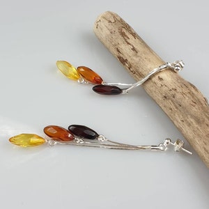 May include: A pair of silver dangle earrings with three amber stones in shades of yellow, orange, and brown. The stones are faceted and have a teardrop shape.