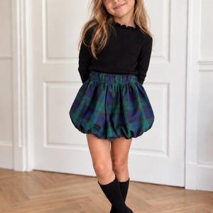 May include: A black long-sleeved ribbed shirt with a ruffled neckline paired with a blue and green plaid bubble skirt, black ribbed knee-high socks, and dark blue velvet Mary Jane shoes with a strap across the top of the foot.