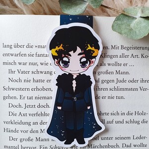 K&ouml;nnte beinhalten: Ein Lesezeichen mit einer Cartoon-Illustration einer Person mit schwarzen Haaren, die einen blau-schwarzen Mantel mit Pelzkragen tr&auml;gt. Der Hintergrund ist ein sternen&uuml;bers&auml;ter Nachthimmel.
