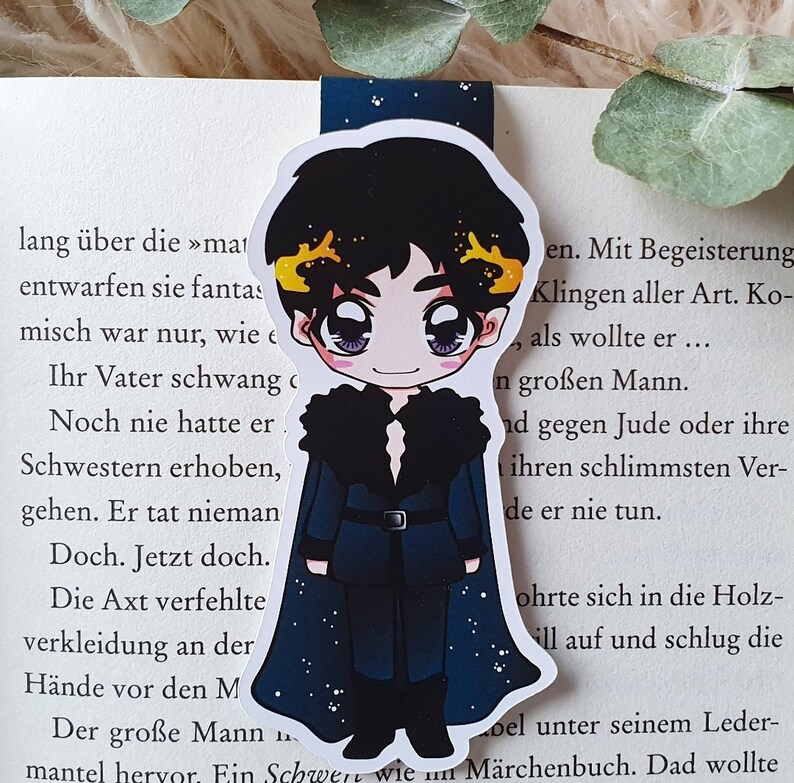 K&ouml;nnte beinhalten: Ein Lesezeichen mit einer Cartoon-Illustration einer Person mit schwarzen Haaren, die einen blau-schwarzen Mantel mit Pelzkragen tr&auml;gt. Der Hintergrund ist ein sternen&uuml;bers&auml;ter Nachthimmel.