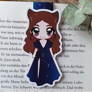 K&ouml;nnte beinhalten: Ein blaues und wei&szlig;es Lesezeichen mit einer Cartoon-Illustration einer Frau mit braunen Haaren, die ein blaues Kleid mit einem wei&szlig;en Sternenmuster tr&auml;gt. Das Lesezeichen hat eine Katzenohrform.