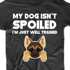May include: Black t-shirt with the text "MY DOG ISN'T SPOILED I'M JUST WELL TRAINED" in white. A cartoon illustration of a German Shepherd dog is featured below the text. The dog has brown and black fur and is peeking over the bottom of the shirt.