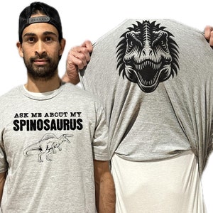 May include: A heather gray t-shirt featuring the black text ASK ME ABOUT MY SPINOSAURUS above a line drawing of a Spinosaurus. The inside of the shirt is pulled up to reveal a large black graphic of a dinosaur head on the back of the fabric.