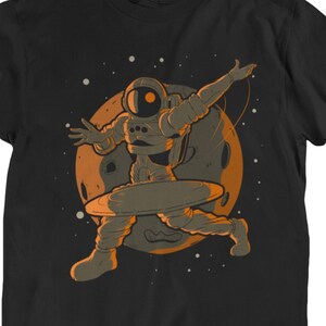 以下が含まれることがあります： 宇宙で惑星の上で踊る宇宙飛行士のオレンジ色のイラストが描かれた黒いTシャツ