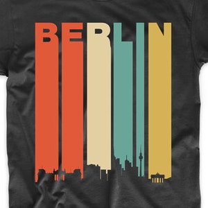 May include: Black t-shirt with a retro-style skyline of Berlin, Germany. The word "BERLIN" is printed in large letters above the skyline, with each letter in a different color.