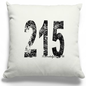 May include: Cream-colored square throw pillow with a large, distressed black number "215" printed on the front. Below the number is the text "Philadelphia, PA" in a smaller, cursive font. The pillow is a decorative home accent.