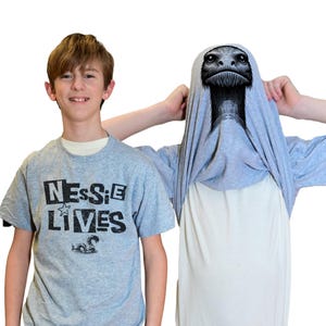May include: A light gray t-shirt featuring the black text "NESSIE LIVES" with a small star and a graphic of a sea creature. A second light gray t-shirt is pulled over a head, displaying a detailed black and white illustration of a creature's face.