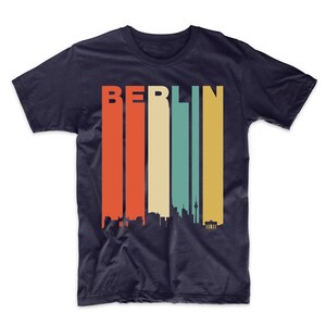 May include: A navy blue t-shirt with a retro-style skyline of Berlin, Germany. The word "BERLIN" is printed in white letters above the skyline. The skyline is made up of various buildings and a tower, all in black.