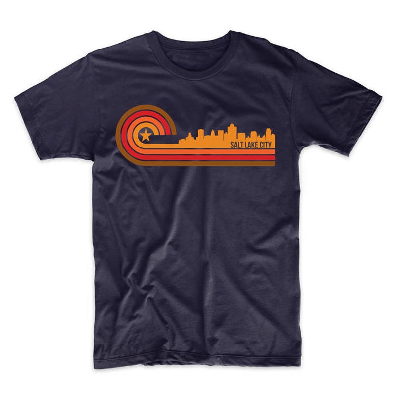 May include: A navy blue t-shirt with a retro-style graphic of the Salt Lake City skyline in orange, red, and yellow. The graphic features a star and stripes.