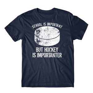Op de afbeelding: Een marineblauw T-shirt met een witte hockeypuck-afbeelding en de tekst "School is belangrijk, maar hockey is belangrijker".