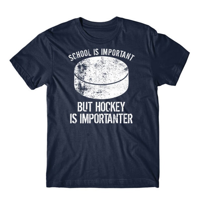 Op de afbeelding: Een marineblauw T-shirt met een witte hockeypuck-afbeelding en de tekst "School is belangrijk, maar hockey is belangrijker".