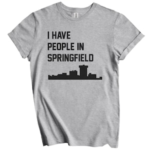 Puede incluir: Camiseta gris claro con mangas enrolladas. La camiseta presenta el texto "I HAVE PEOPLE IN SPRINGFIELD" en negrita, en negro, sobre un gráfico de horizonte negro. La camiseta está hecha de un material suave y cómodo.