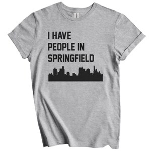 Puede incluir: Camiseta gris claro con el texto negro "I HAVE PEOPLE IN SPRINGFIELD" sobre un gráfico de horizonte negro. La camiseta tiene las mangas enrolladas.