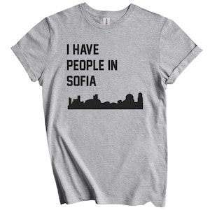 May include: A light gray t-shirt with rolled-up sleeves. The shirt features the text "I HAVE PEOPLE IN SOFIA" in black, above a black silhouette of the Sofia skyline.