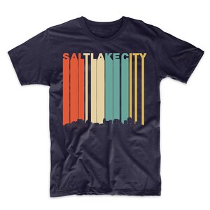 May include: A navy blue t-shirt with a retro-style graphic of the Salt Lake City skyline. The graphic features a multi-colored striped background with the city name in white letters.