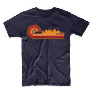 Puede incluir: Una camiseta azul marino con un gr&aacute;fico de estilo retro del horizonte de Los &Aacute;ngeles en naranja, rojo y marr&oacute;n. El gr&aacute;fico presenta una estrella y el texto "LOS ANGELES".