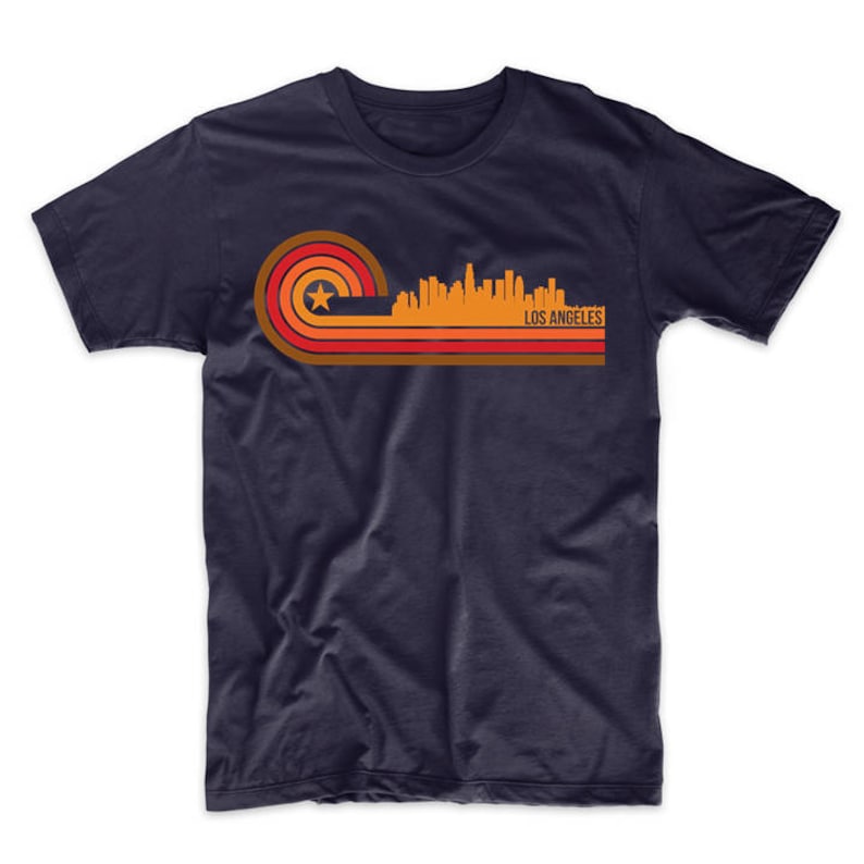 Puede incluir: Una camiseta azul marino con un gr&aacute;fico de estilo retro del horizonte de Los &Aacute;ngeles en naranja, rojo y marr&oacute;n. El gr&aacute;fico presenta una estrella y el texto "LOS ANGELES".