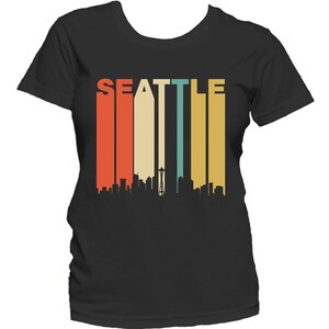 May include: A black t-shirt with a retro-style graphic of the Seattle skyline. The graphic features a colorful striped background with the word "SEATTLE" in white letters.