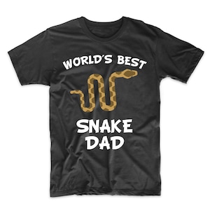 Pode incluir: Camiseta preta com texto branco que diz "World's Best Snake Dad" e uma imagem de desenho animado de uma cobra marrom.