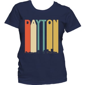 May include: A navy blue t-shirt with a retro-style graphic of the word "DAYTON" in colorful vertical stripes, with a skyline silhouette below.