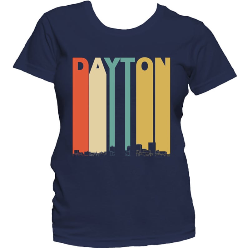 May include: A navy blue t-shirt with a retro-style graphic of the word "DAYTON" in colorful vertical stripes, with a skyline silhouette below.