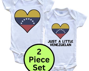 Puede incluir: Un conjunto de dos bodies blancos de manga corta para bebé. Uno presenta un gráfico de corazón con los colores amarillo, azul y rojo de la bandera venezolana y estrellas blancas. El otro incluye el mismo corazón y el texto JUST A LITTLE VENEZUELAN.