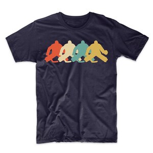 Pode incluir: Uma camiseta azul marinho com um gr&aacute;fico de estilo retro de quatro goleiros de h&oacute;quei em fila. Os goleiros est&atilde;o em silhueta vermelha, laranja, verde e azul.