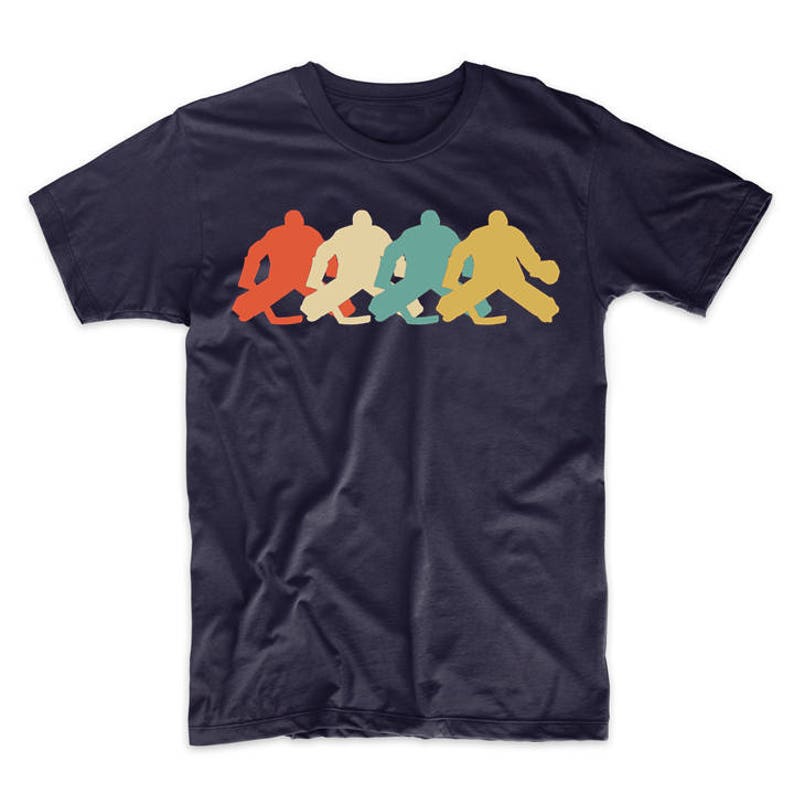 Pode incluir: Uma camiseta azul marinho com um gr&aacute;fico de estilo retro de quatro goleiros de h&oacute;quei em fila. Os goleiros est&atilde;o em silhueta vermelha, laranja, verde e azul.