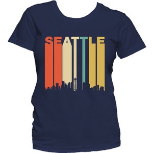 May include: A navy blue t-shirt with a retro-style graphic of the Seattle skyline and the word "SEATTLE" in colorful vertical stripes.