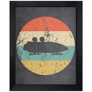 May include: A framed wall art print featuring a black silhouette of a two-person bobsled with riders inside. The bobsled is centered over a circular background with horizontal stripes in orange, cream, teal, and yellow, all with a distressed texture.