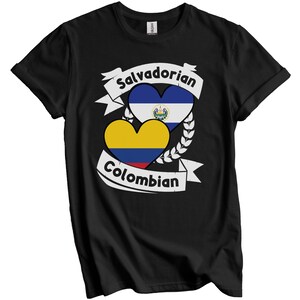 Op de afbeelding: Een zwart T-shirt met korte mouwen en een grafisch ontwerp met twee hartvormige vlaggen, die El Salvador en Colombia vertegenwoordigen. Het ontwerp bevat witte banners met de tekst Salvadorian en Colombian.